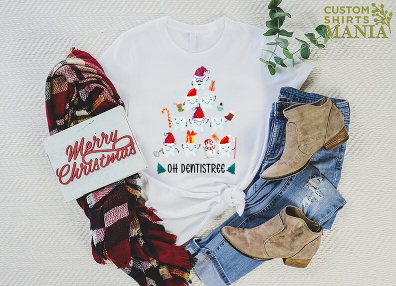 以下が含まれることがあります： サンタの帽子をかぶった歯でできたクリスマスツリーのデザインが施された白いTシャツ。ツリーの下には「Oh Dentistree」という文字があります。