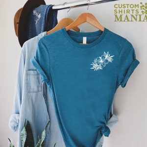 May include: A teal blue t-shirt with a white floral design on the left chest. The shirt is hanging on a wooden hanger. The text "CUSTOM SHIRTS MANIA" is visible in the top right corner of the image.
