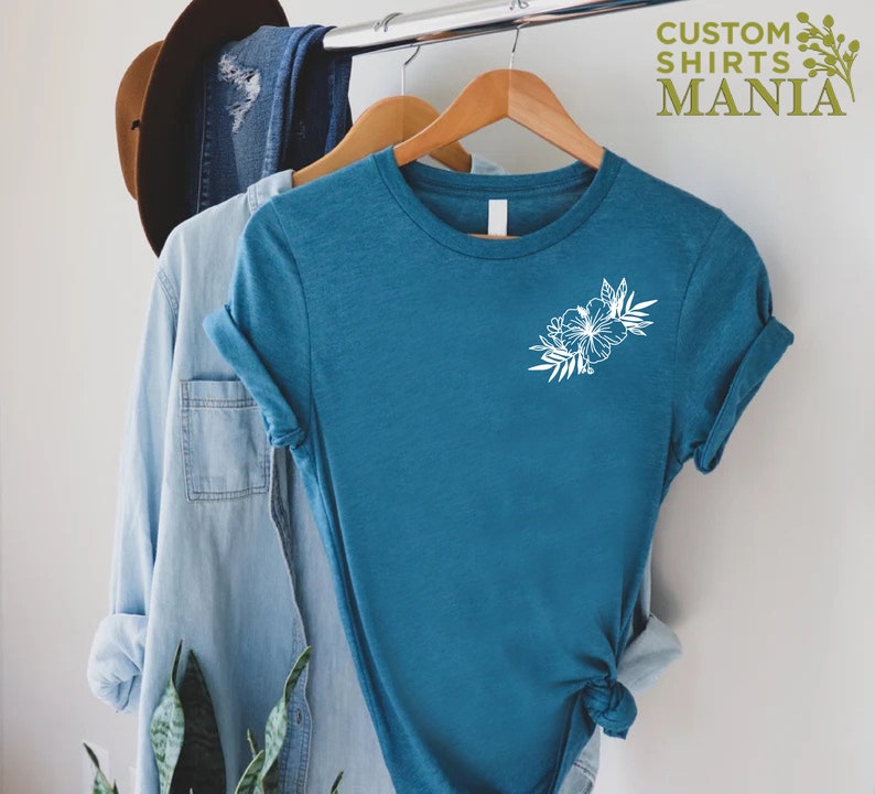 May include: A teal blue t-shirt with a white floral design on the left chest. The shirt is hanging on a wooden hanger. The text "CUSTOM SHIRTS MANIA" is visible in the top right corner of the image.