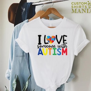 K&ouml;nnte beinhalten: Wei&szlig;es T-Shirt mit einem bunten Puzzle-Herz und dem Text "Ich liebe jemanden mit AUTISMUS" in Regenbogenfarben.