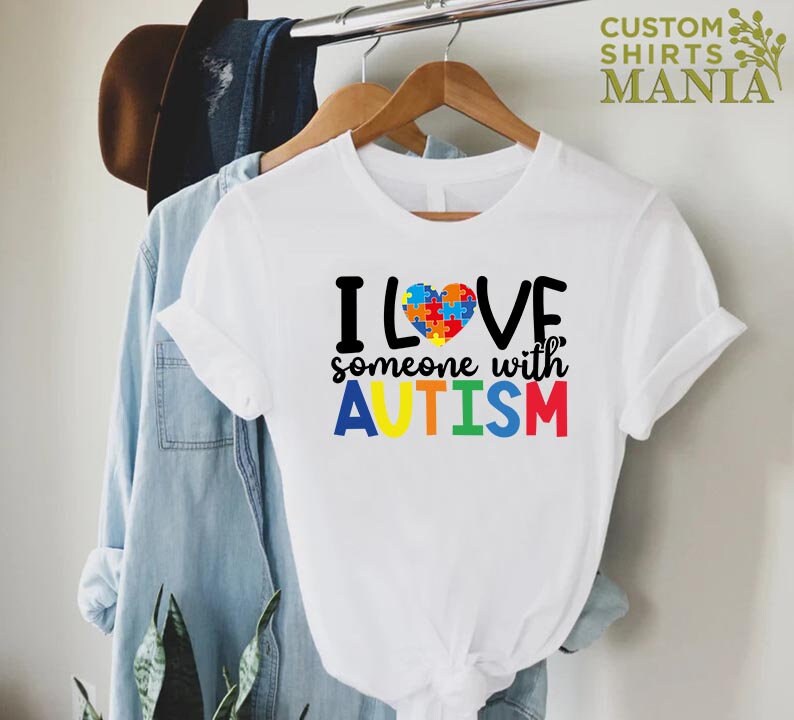 K&ouml;nnte beinhalten: Wei&szlig;es T-Shirt mit einem bunten Puzzle-Herz und dem Text "Ich liebe jemanden mit AUTISMUS" in Regenbogenfarben.