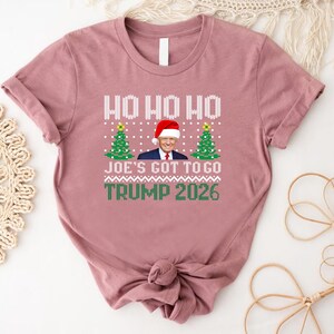 Op de afbeelding: Een mauve t-shirt met korte mouwen en een grafische print met de tekst HO HO HO, JOE'S GOT TO GO en TRUMP 2026. Het ontwerp bevat een centrale afbeelding van Donald Trump met een kerstmuts, geflankeerd door twee groene kerstbomen.