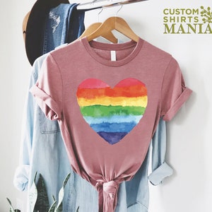 Peut inclure: Un t-shirt rose poussi&eacute;reux avec un motif de c&oelig;ur arc-en-ciel. Le c&oelig;ur est divis&eacute; en bandes horizontales rouges, oranges, jaunes, vertes, bleues et violettes. Le t-shirt est suspendu &agrave; un cintre en bois avec d'autres v&ecirc;tements. Le texte "CUSTOM SHIRTS MANIA" est visible.