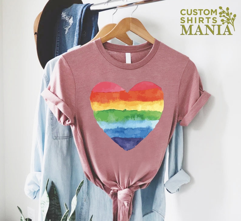 Peut inclure: Un t-shirt rose poussi&eacute;reux avec un motif de c&oelig;ur arc-en-ciel. Le c&oelig;ur est divis&eacute; en bandes horizontales rouges, oranges, jaunes, vertes, bleues et violettes. Le t-shirt est suspendu &agrave; un cintre en bois avec d'autres v&ecirc;tements. Le texte "CUSTOM SHIRTS MANIA" est visible.