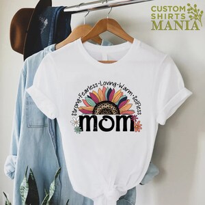 May include: White t-shirt featuring a colourful sunflower graphic and the word "MOM" in black text. The sunflower has a leopard print centre and is surrounded by the words "Strong, Fearless, Loving, Warm, Selfless".