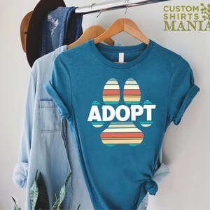 May include: Teal blue t-shirt with a retro-style rainbow paw print graphic and the word "ADOPT" in white text.