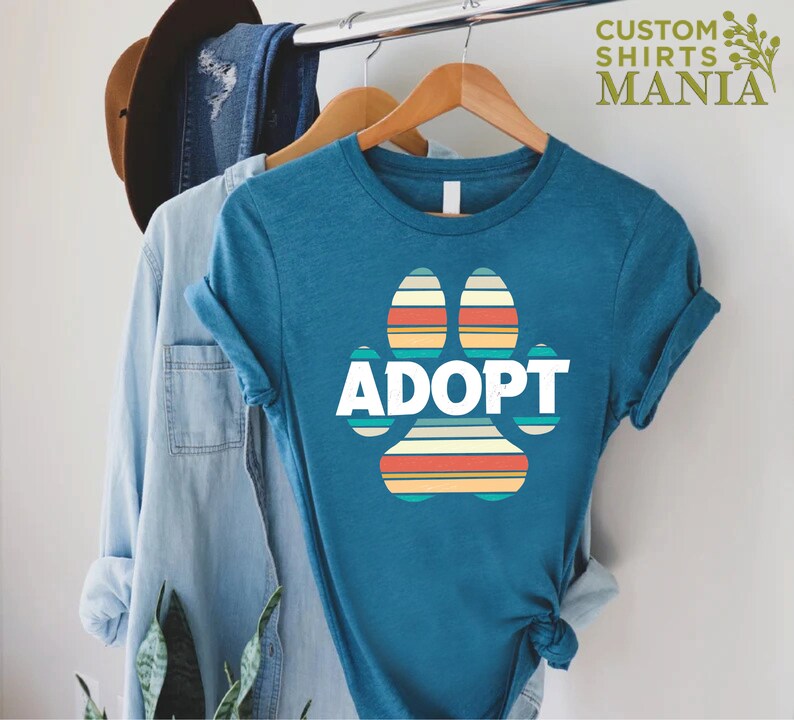 May include: Teal blue t-shirt with a retro-style rainbow paw print graphic and the word "ADOPT" in white text.
