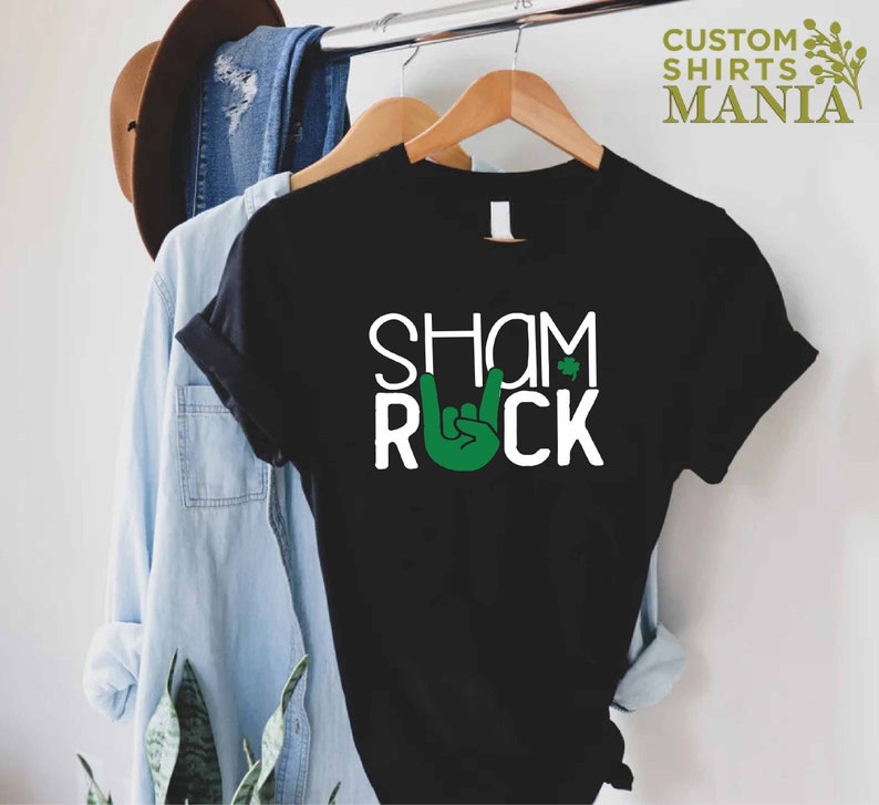 Peut inclure: T-shirt noir avec du texte blanc qui dit "Shamrock" et "Rock" avec une main verte faisant le signe rock and roll.