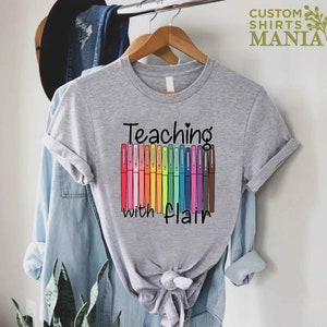 May include: A gray t-shirt with the text "Teaching with flair" and a row of colorful markers.