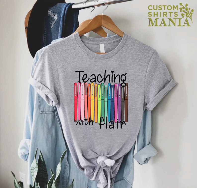 May include: A gray t-shirt with the text "Teaching with flair" and a row of colorful markers.