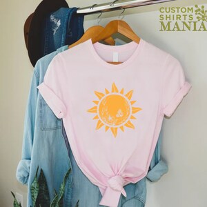 Puede incluir: Una camiseta rosa con un gr&aacute;fico de sol amarillo que presenta un dise&ntilde;o floral blanco y mariposas. El gr&aacute;fico del sol est&aacute; centrado en la camiseta.