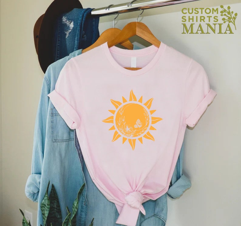 Puede incluir: Una camiseta rosa con un gr&aacute;fico de sol amarillo que presenta un dise&ntilde;o floral blanco y mariposas. El gr&aacute;fico del sol est&aacute; centrado en la camiseta.