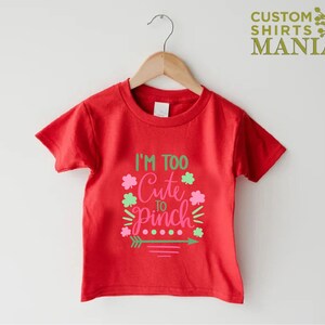 May include: Red short-sleeved t-shirt with the text "I'm Too Cute To Pinch" in pink and green lettering with shamrock designs.
