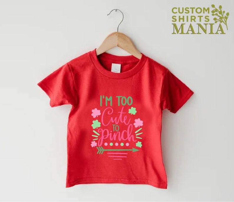 May include: Red short-sleeved t-shirt with the text "I'm Too Cute To Pinch" in pink and green lettering with shamrock designs.