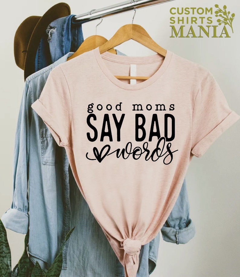 May include: A pink t-shirt with the text "good moms say bad words" in black lettering. The text is in a cursive font with a heart symbol before the word "words".