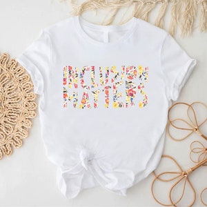 Puede incluir: Camiseta blanca con el texto "Inclusion Matters" en estampado floral.