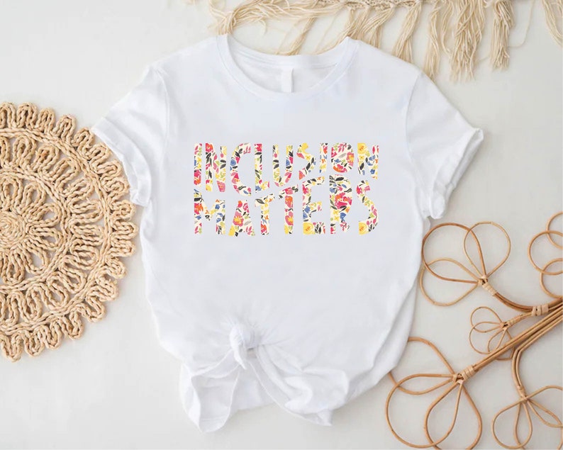 Puede incluir: Camiseta blanca con el texto "Inclusion Matters" en estampado floral.