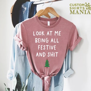 May include: A pink t-shirt with the text "Look at me being all festive and shit" in white lettering and a small green Christmas tree with lights below the text.