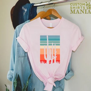 May include: A pink t-shirt with a white silhouette of three cacti in front of a sunset-colored striped background. The text "CUSTOM SHIRTS MANIA" is in the top right corner of the image.