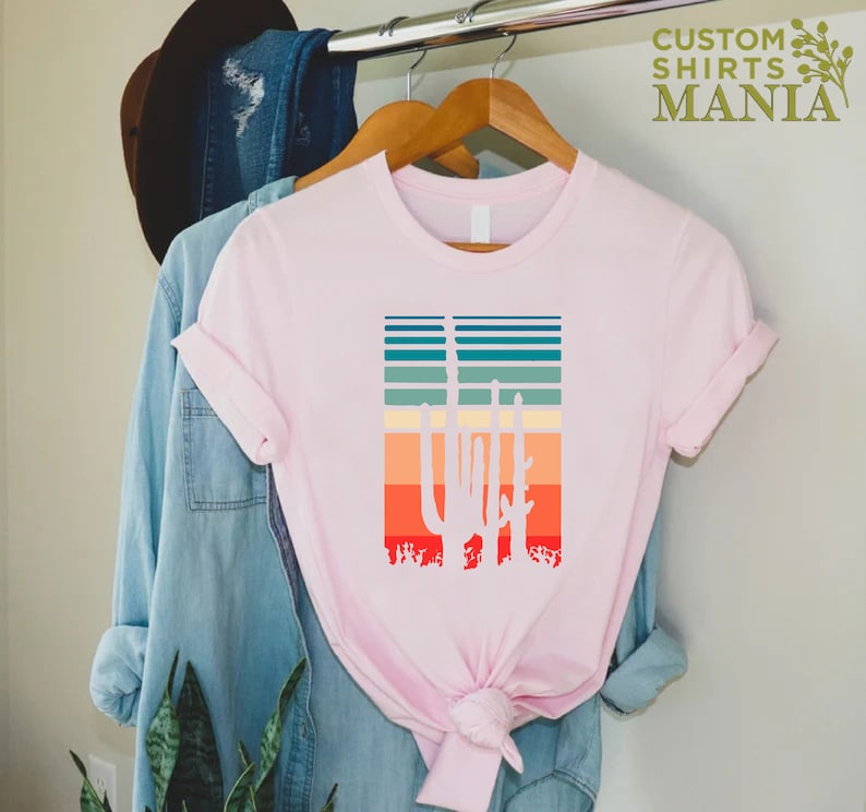 May include: A pink t-shirt with a white silhouette of three cacti in front of a sunset-colored striped background. The text "CUSTOM SHIRTS MANIA" is in the top right corner of the image.