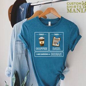 May include: A teal blue t-shirt with a graphic design of a coffee cup and a clipboard with the text "First - Coffee, Then - Data, I am earning a: Break".