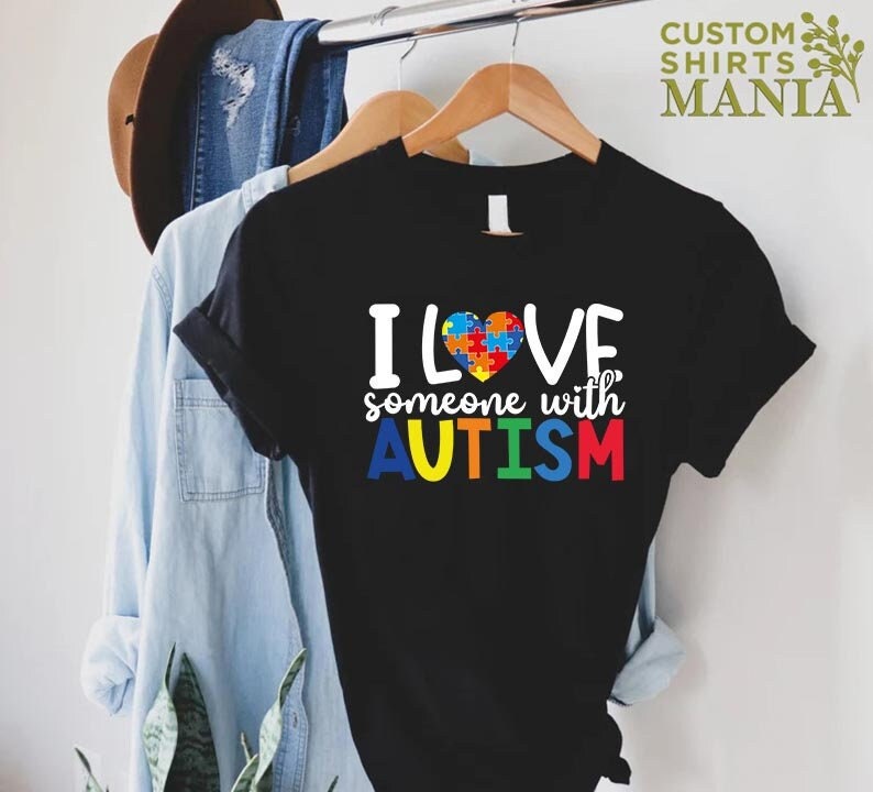 K&ouml;nnte beinhalten: Schwarzes T-Shirt mit dem Text "ICH LIEBE jemanden mit AUTISMUS" in Regenbogenfarben. Ein Puzzle-Herz befindet sich in der Mitte des Textes.