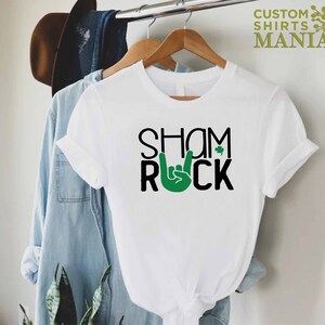 Peut inclure: T-shirt blanc avec du texte noir qui dit "Shamrock" avec un tr&egrave;fle vert et une main verte faisant le signe rock and roll.