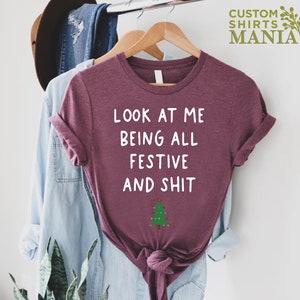 May include: A maroon t-shirt with the text "Look at me being all festive and shit" in white lettering and a small green Christmas tree graphic.