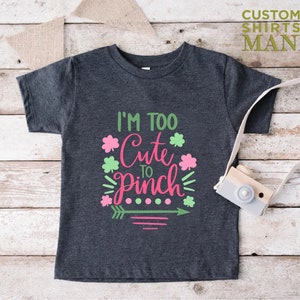 May include: A dark grey t-shirt with a green and pink graphic that says "I'm Too Cute To Pinch" with shamrock designs and an arrow.