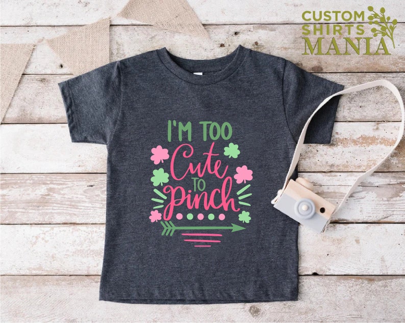 May include: A dark grey t-shirt with a green and pink graphic that says "I'm Too Cute To Pinch" with shamrock designs and an arrow.