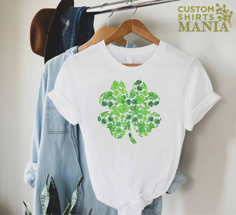 May include: White t-shirt with a green four-leaf clover design made of smaller clovers. The design is centered on the shirt.