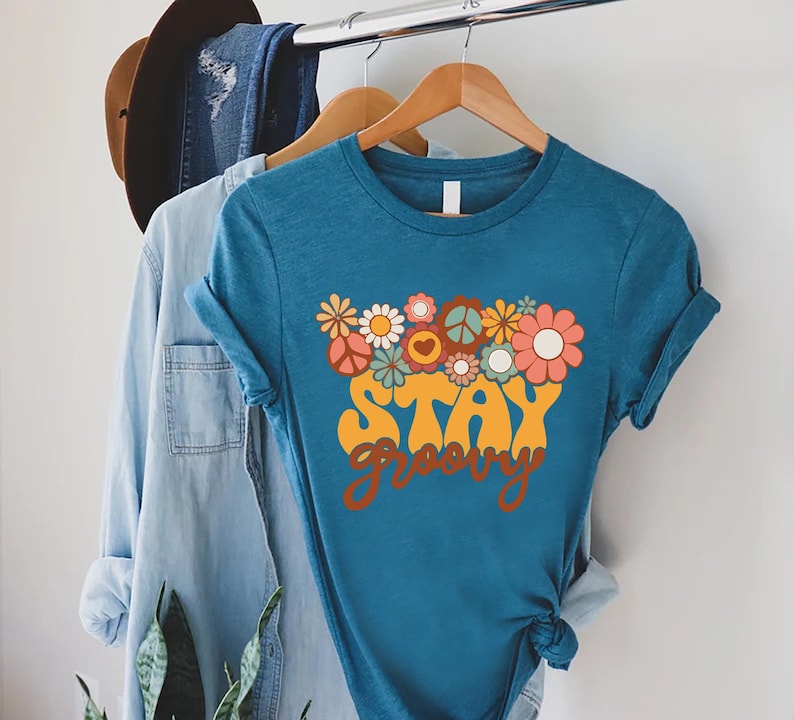 May include: A teal blue t-shirt with a retro-style graphic design featuring the text "Stay Groovy" surrounded by colorful flowers and peace signs.