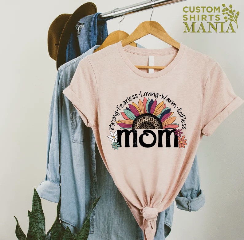 May include: A pink t-shirt featuring a sunflower graphic and the word "MOM" in black. The sunflower has a leopard print centre and is surrounded by the words "Strong, Fearless, Loving, Warm, Selfless".