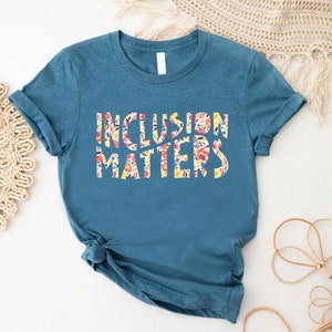 Puede incluir: Una camiseta azul turquesa con las palabras "Inclusion Matters" impresas en blanco con un patr&oacute;n floral.