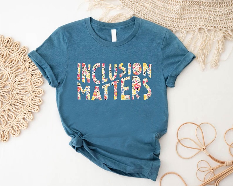 Puede incluir: Una camiseta azul turquesa con las palabras "Inclusion Matters" impresas en blanco con un patr&oacute;n floral.