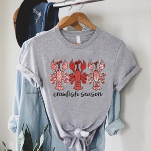 May include: A light gray t-shirt featuring three red crawfish with decorative patterns. The middle crawfish is solid red, while the outer two have pink polka dots. The words "crawfish season" are written in black script below the crawfish.
