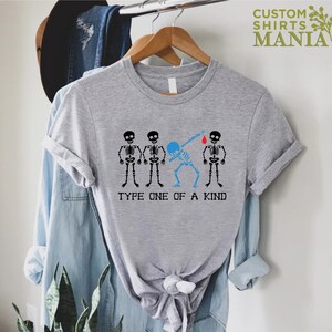 以下が含まれることがあります： 4 つの骸骨のグラフィックが描かれたグレーのTシャツ。1 つの骸骨が指に赤い血の滴をつけた状態でダブをしています。「Type One of a Kind」というテキストが骸骨の下にあります。