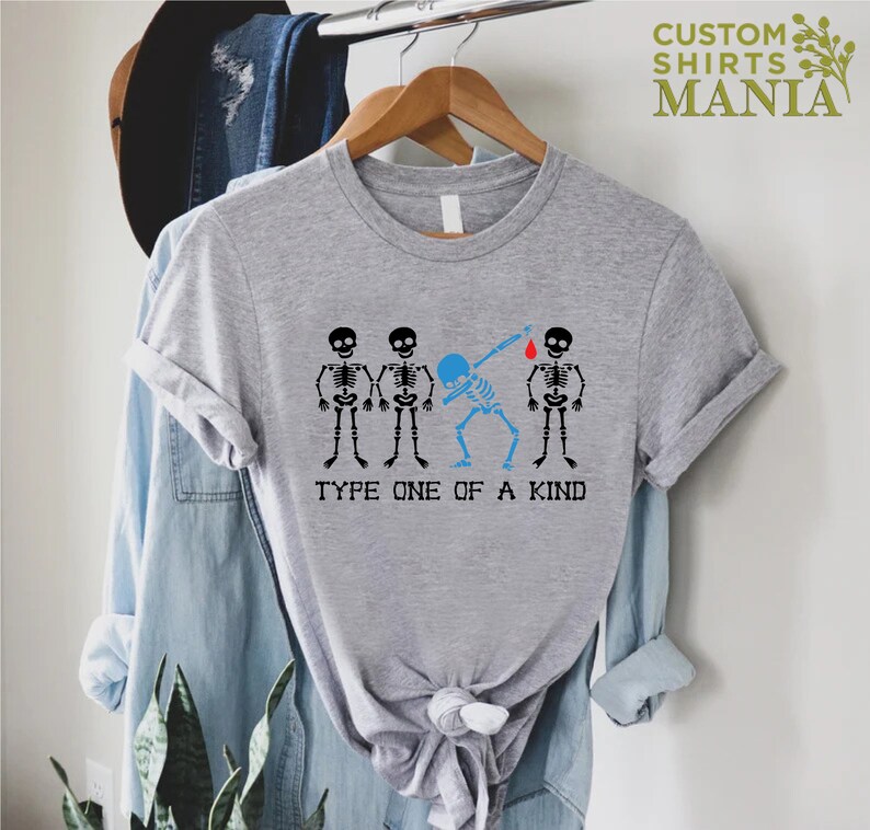 以下が含まれることがあります： 4 つの骸骨のグラフィックが描かれたグレーのTシャツ。1 つの骸骨が指に赤い血の滴をつけた状態でダブをしています。「Type One of a Kind」というテキストが骸骨の下にあります。