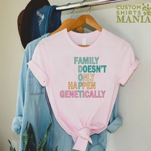 Op de afbeelding: Roze T-shirt met de tekst "FAMILY DOESN'T ONLY HAPPEN GENETICALLY" in kleurrijke letters.