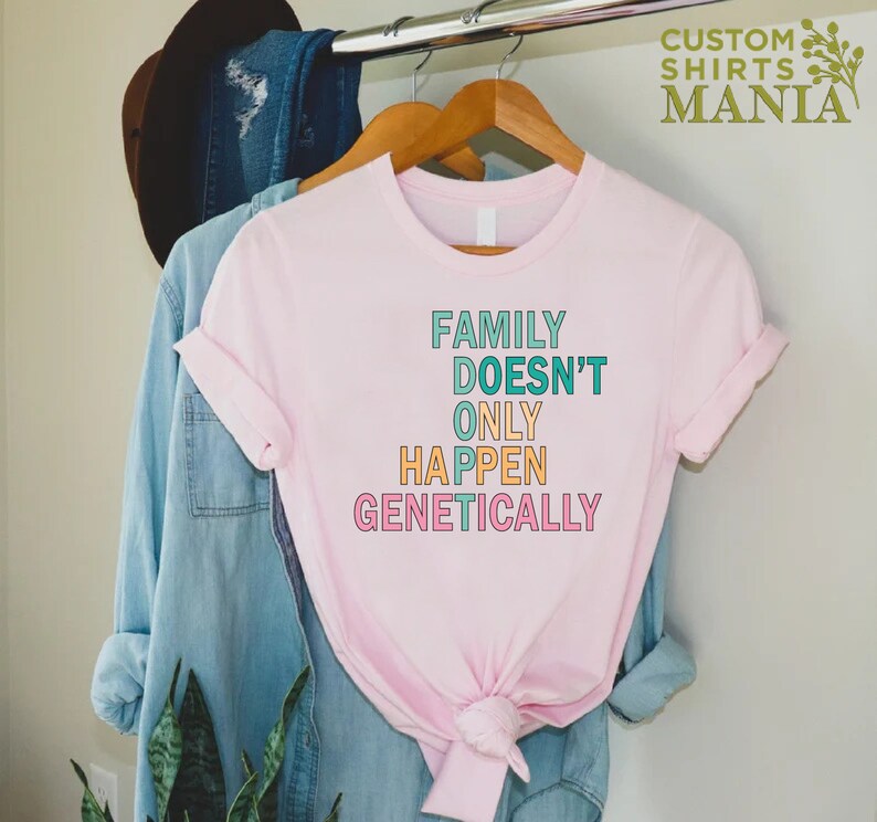 Op de afbeelding: Roze T-shirt met de tekst "FAMILY DOESN'T ONLY HAPPEN GENETICALLY" in kleurrijke letters.