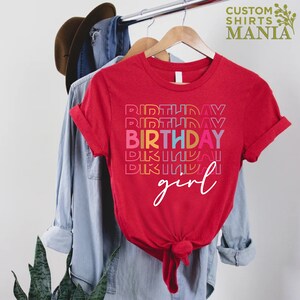 May include: Red t-shirt with the text "BIRTHDAY girl" in rainbow colours. The word "BIRTHDAY" is repeated multiple times in a stacked design.