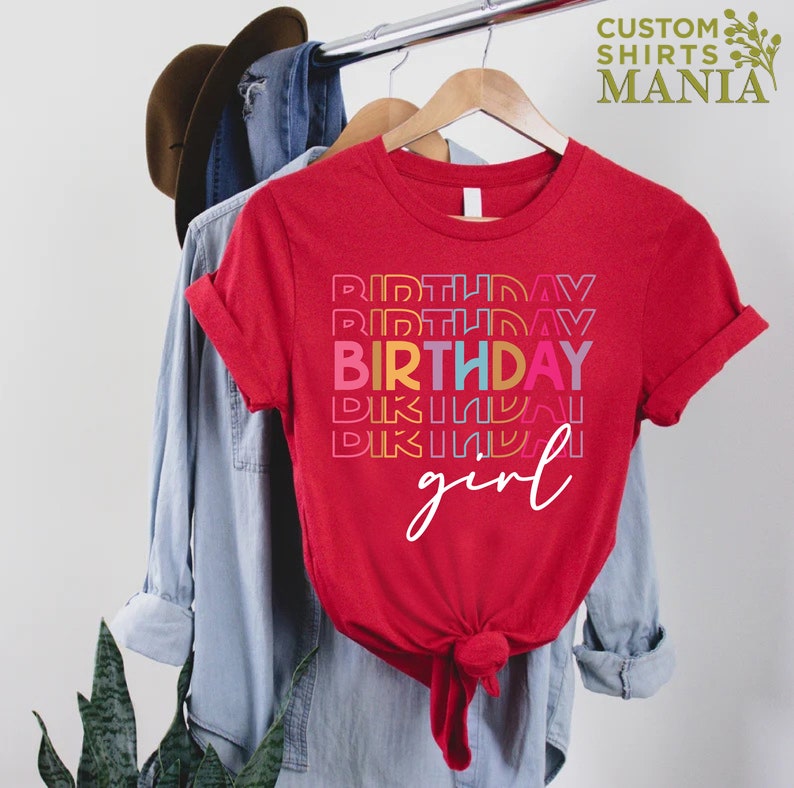 May include: Red t-shirt with the text "BIRTHDAY girl" in rainbow colours. The word "BIRTHDAY" is repeated multiple times in a stacked design.