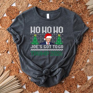 Op de afbeelding: Een donkergrijs gem&ecirc;leerd T-shirt met korte mouwen, voorzien van een wit breipatroon, de tekst HO HO HO, een afbeelding van Donald Trump met een kerstmuts tussen twee groene kerstbomen, de tekst JOE'S GOT TO GO en TRUMP 2024.