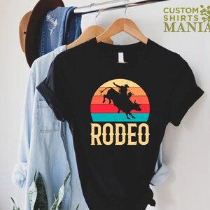 May include: Black t-shirt with a vintage-style sunset graphic of a cowboy riding a bull. The text "RODEO" is printed below the graphic.