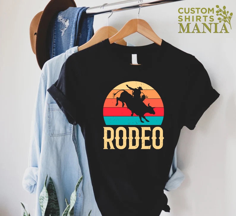 May include: Black t-shirt with a vintage-style sunset graphic of a cowboy riding a bull. The text "RODEO" is printed below the graphic.
