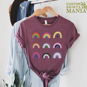 K&ouml;nnte beinhalten: Ein maronfarbenes T-Shirt mit einem Design aus neun verschiedenen bunten Regenb&ouml;gen. Die Regenb&ouml;gen sind in drei Reihen zu je drei angeordnet. Der Text "CUSTOM SHIRTS MANIA" befindet sich in der oberen rechten Ecke des Bildes.