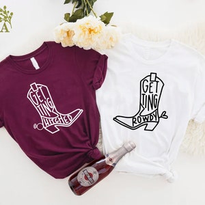 May include: Two t-shirts with a boot design and text. The maroon shirt says "Getting Hitched" and the white shirt says "Getting Rowdy".