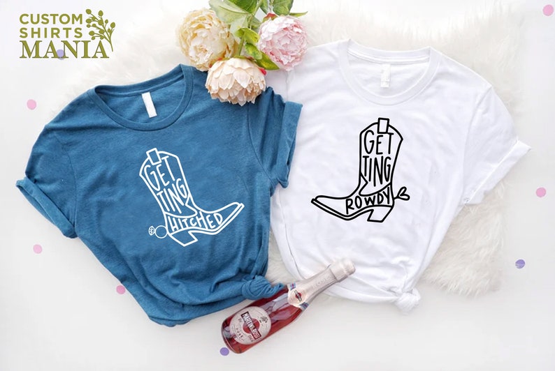 May include: Two t-shirts with a white cowboy boot graphic. The text on the blue shirt says "Getting Hitched" and the text on the white shirt says "Getting Rowdy".