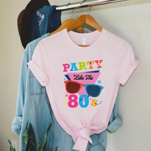 K&ouml;nnte beinhalten: Rosa T-Shirt mit einem Grafikdesign mit dem Text "Party Like The 80s" in bunten Buchstaben und einer Sonnenbrille im Retro-Design.