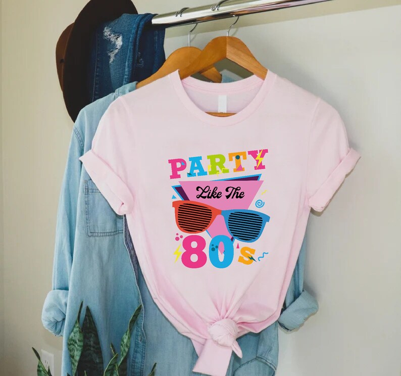 K&ouml;nnte beinhalten: Rosa T-Shirt mit einem Grafikdesign mit dem Text "Party Like The 80s" in bunten Buchstaben und einer Sonnenbrille im Retro-Design.